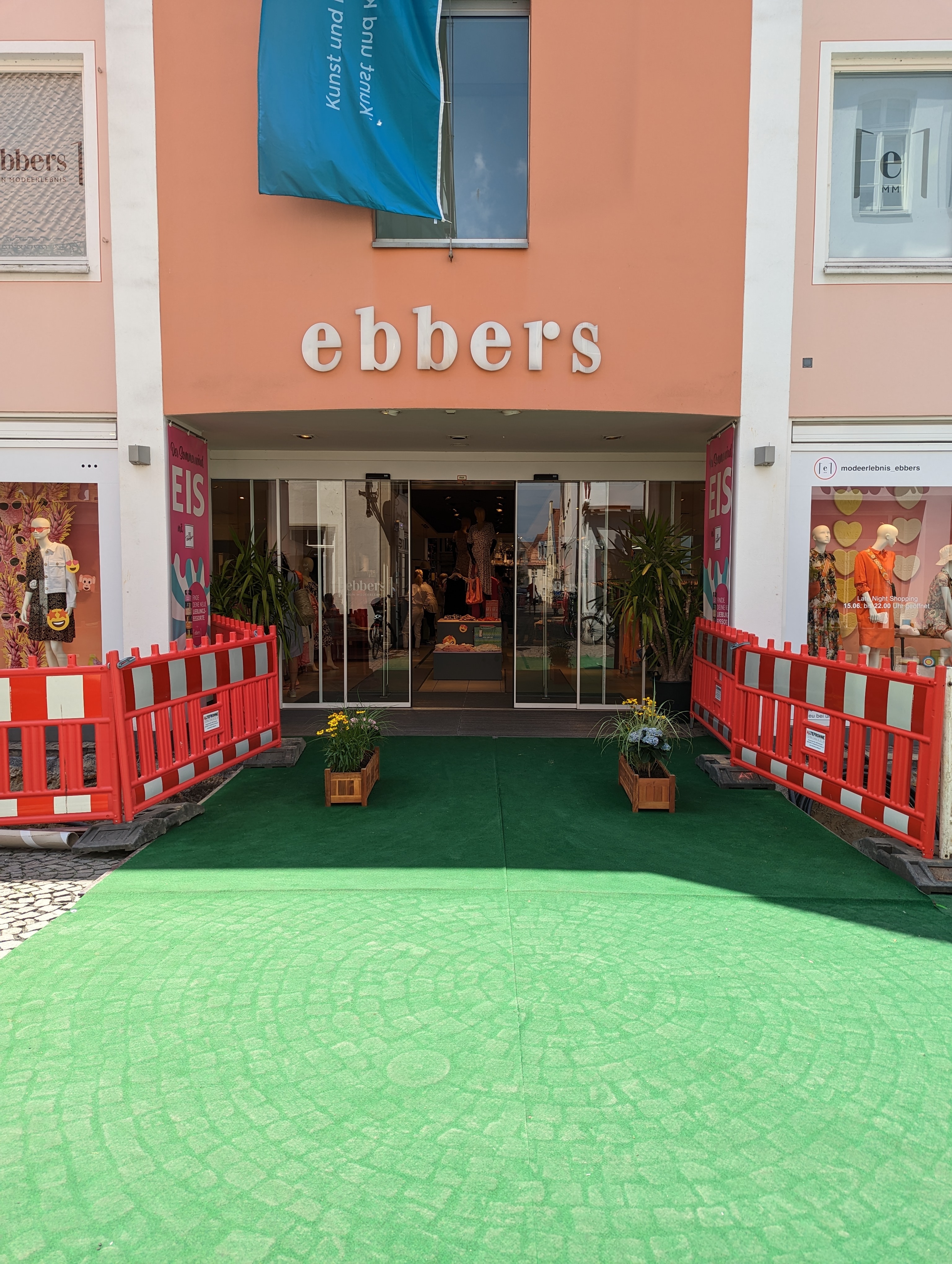 Zugang zu einem Modegeschäft von der Fußgängerzone aus, davor ein grüner Teppich und Blumenkästen. Absperrungen links und rechts bilden ein "Tor".