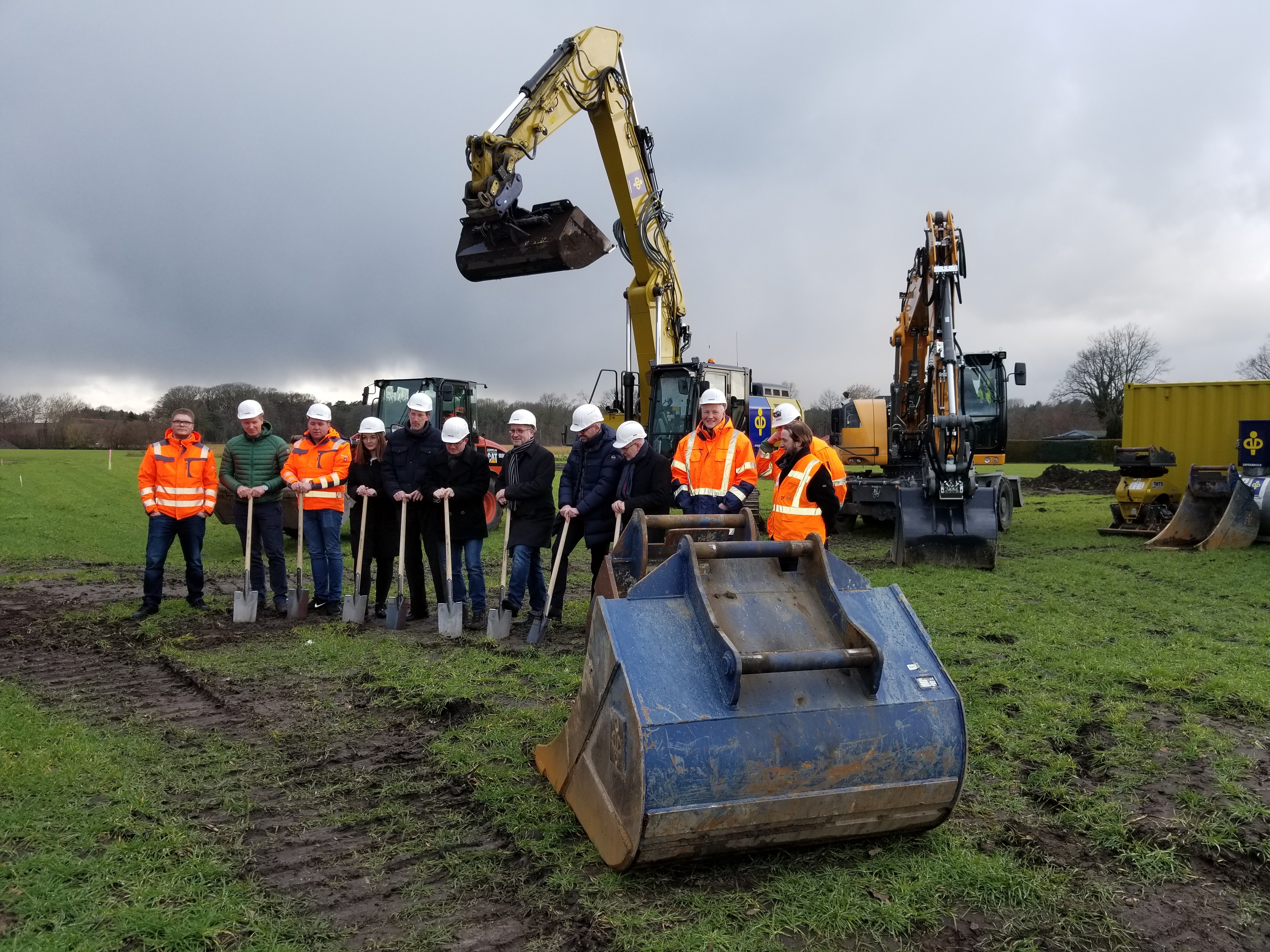 Aufstellung von Menschen und Maschinen für den ersten Spatenstich. Die Vertreter von verschiedenen Projektbeteiligten tragen Helme und haben Spaten in der Hand. 
